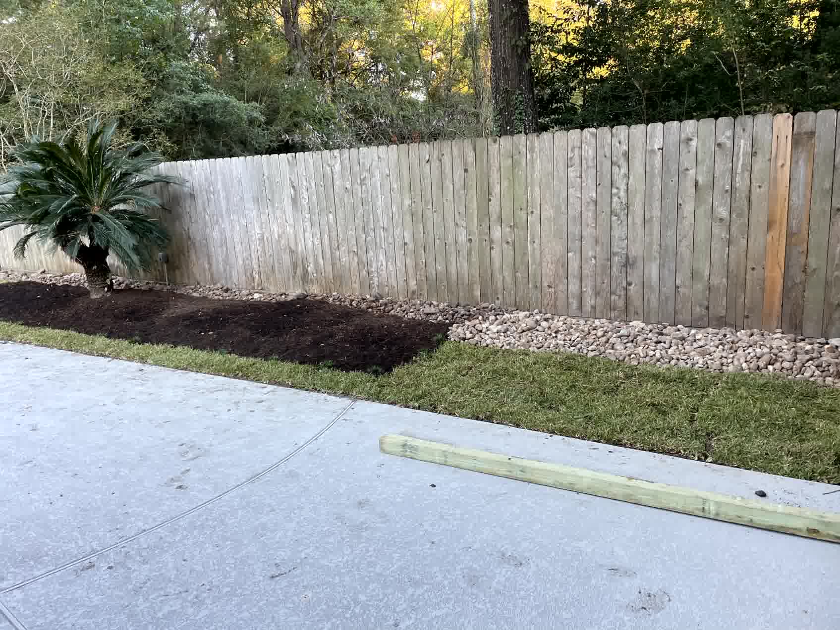 Curved raised flower bed with black mulch and border stones surrounds tree in suburban yard with green grass and wooden fence.