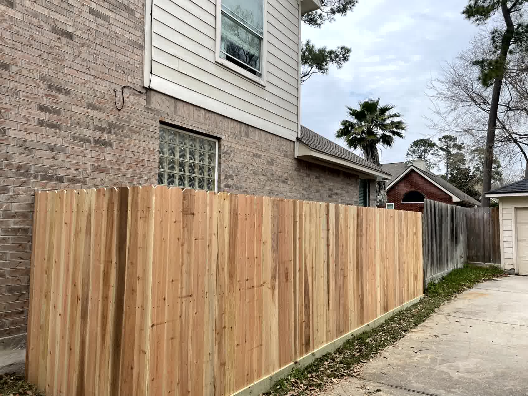 Newly installed cedar privacy fence borders brick house and concrete driveway, offering backyard security and curb appeal.